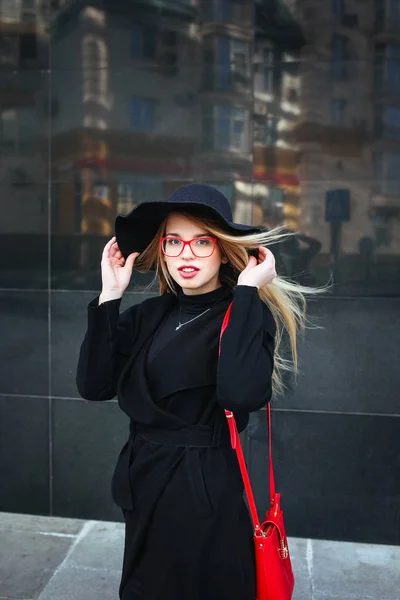 Hermosa joven con un abrigo y sombrero negro, con gafas rojas y una bolsa roja caminando por la ciudad. Reflejo de la ciudad en la ventana —  Fotos de Stock