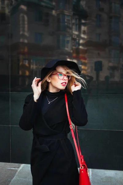 Bella giovane donna in un cappotto nero e cappello, indossando occhiali rossi e una borsa rossa che cammina in città. Riflessione della città nella finestra — Foto Stock