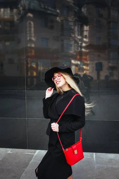 Красива молода жінка в чорному пальто і капелюсі, одягнена в червоні окуляри і червона сумка, що йде в місті. Відображення міста у вікні — стокове фото
