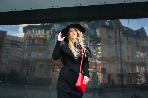 Piękna młoda kobieta w czarny płaszcz i czapkę, czerwone okulary i czerwony worek spaceru po mieście. Odbicie miasta w oknie — Zdjęcie stockowe