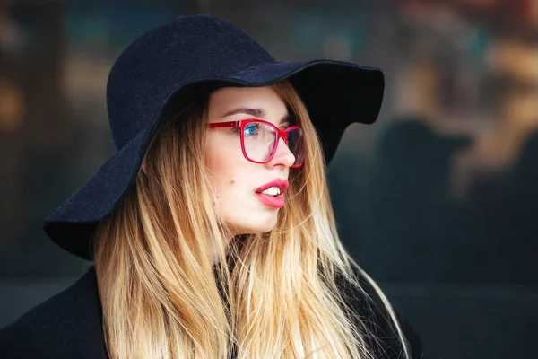 Όμορφη νεαρή γυναίκα σε ένα μαύρο παλτό και καπέλο, φοράει κόκκινα γυαλιά και μια κόκκινη τσάντα το περπάτημα στην πόλη. Αντανάκλαση της πόλης στο παράθυρο — Φωτογραφία Αρχείου