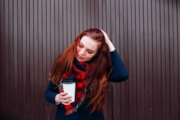 Hermosa joven feliz sosteniendo una taza de papel y tomando café. Mujer alegre con el pelo largo rojo en la calle bebiendo café de la mañana — Foto de Stock