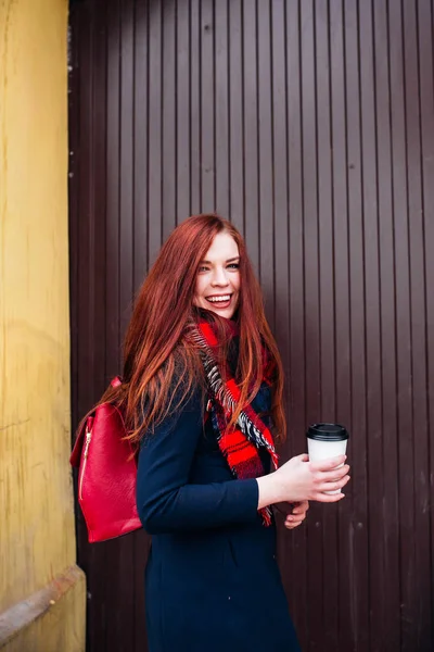 Прекрасная счастливая молодая женщина держит бумажный стаканчик и пьет кофе. Веселая женщина с рыжими длинными волосами на улице пьет утренний кофе — стоковое фото