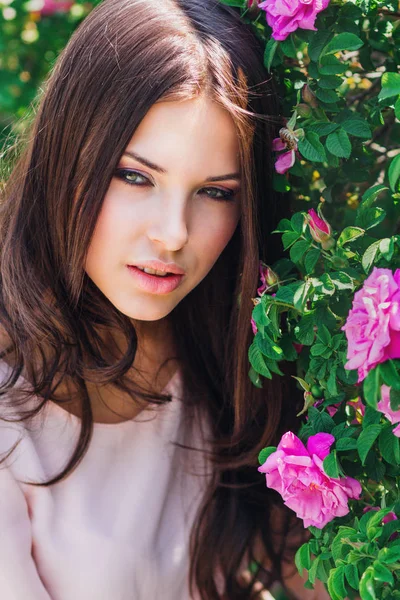 Όμορφη νεαρή γυναίκα με μακριά σγουρά μαλλιά ποζάρει κοντά τριαντάφυλλα μέσα σε έναν κήπο. Η έννοια της διαφημίσεως άρωμα. — Φωτογραφία Αρχείου