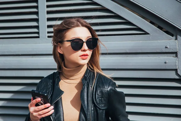 Mode portret mooie vrouw in zwarte rock stijl in zonnebril met mobiele telefoon via de grijze achtergrond in stad — Stockfoto
