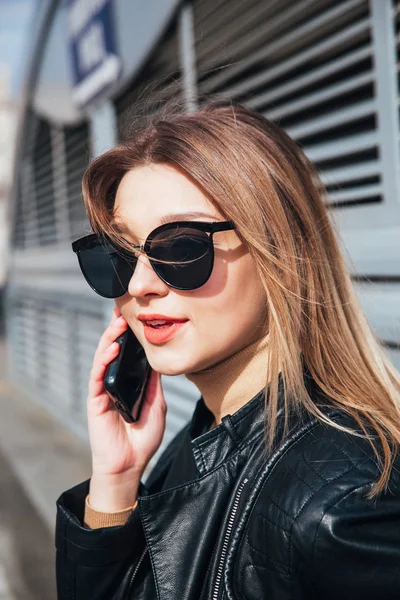 Modeporträt hübsche Frau im schwarzen Rock-Stil in Sonnenbrille mit Handy vor grauem Hintergrund in der Stadt — Stockfoto