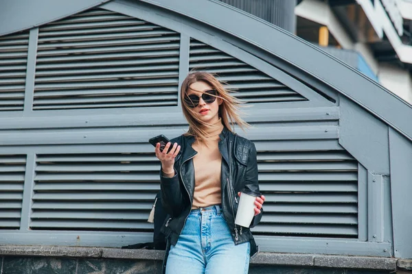Модный портрет симпатичной женщины в черном стиле рок в солнцезащитных очках с мобильным телефоном на заднем плане в городе. — стоковое фото