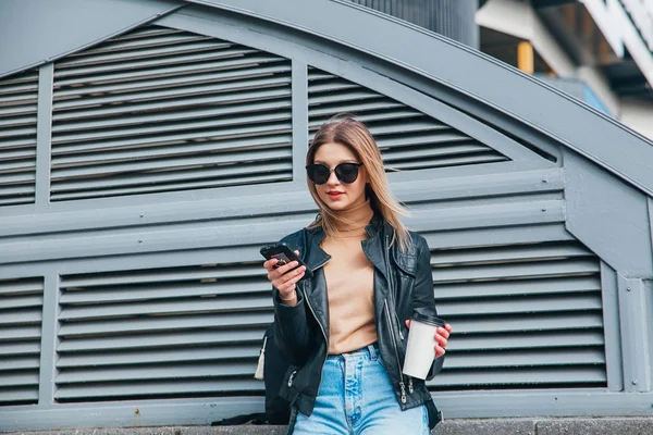 Retrato de moda mujer bonita en estilo rock negro en gafas de sol utilizando el teléfono móvil sobre fondo gris en la ciudad .Girl con cofee — Foto de Stock