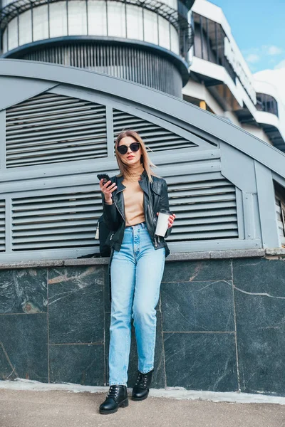 Fashion portrait pretty woman in black rock style in sunglasses using mobile phone over gray background in city .Girl with cofee