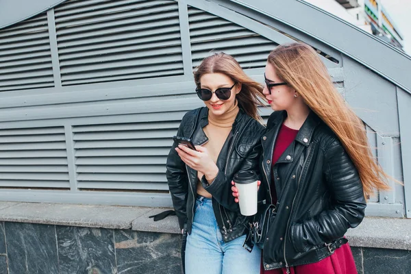 Two Women Talking in the City.Outdoor lifestyle portrait of two best friends hipster girls wearing stylish Leather Jacket and sunglasses with cofee, going crazy and having great time together