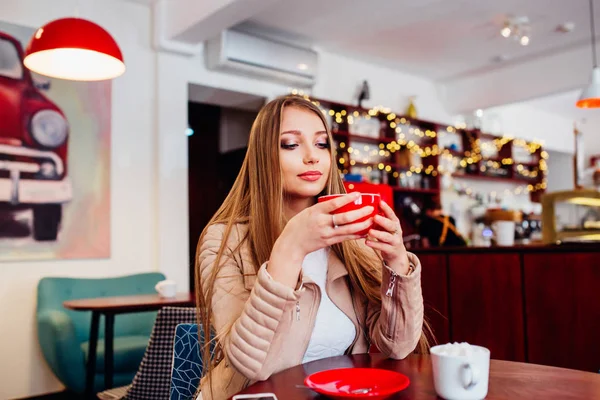 Портрет молодой великолепной женщины, пьющей кофе и выглядывающей с улыбкой из окна кофейни, наслаждаясь отдыхом, приятным обедом деловой женщины в современном кафе во время ее рабочего перерыва — стоковое фото