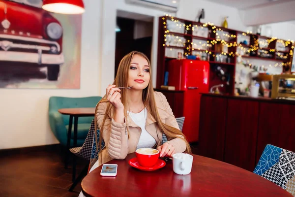 Портрет молодой великолепной женщины, пьющей кофе и выглядывающей с улыбкой из окна кофейни, наслаждаясь отдыхом, приятным обедом деловой женщины в современном кафе во время ее рабочего перерыва — стоковое фото