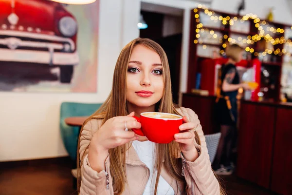 Hermosa joven con una taza de café rojo en un café. Mujer bebiendo café con leche caliente en la acogedora cafetería — Foto de Stock