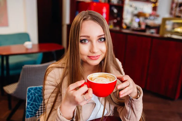 Hermosa joven con una taza de café rojo en un café. Mujer bebiendo café con leche caliente en la acogedora cafetería — Foto de Stock