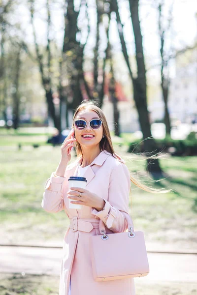 Mujer de moda con teléfono y cofee en la ciudad. Mujer de moda en gafas de sol y chaqueta rosa al aire libre — Foto de Stock