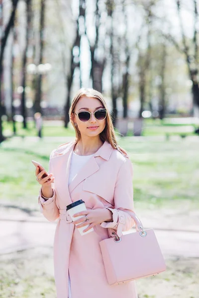 Mujer de moda con teléfono y cofee en la ciudad. Mujer de moda en gafas de sol y chaqueta rosa al aire libre — Foto de Stock