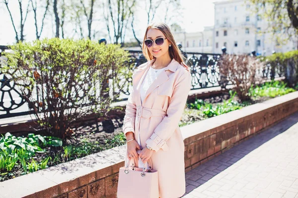 Mooie stijlvolle meisje lopen en poseren in witte jurk en roze jas in de stad. Buiten zomer portret van stijlvolle jongedame in zonnebril — Stockfoto