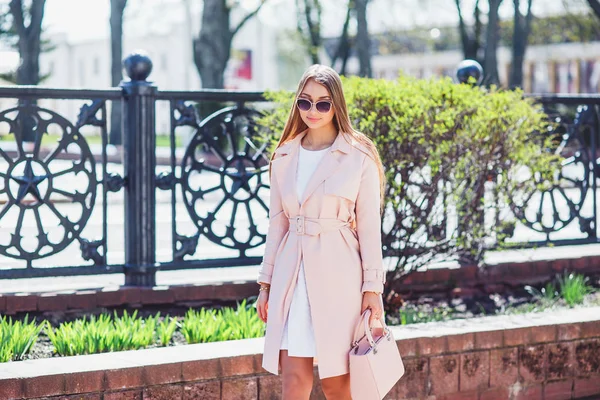 Giovane bella ragazza elegante a piedi e in posa in abito bianco e cappotto rosa in città. Ritratto estivo all'aperto di giovane donna di classe in occhiali da sole — Foto Stock