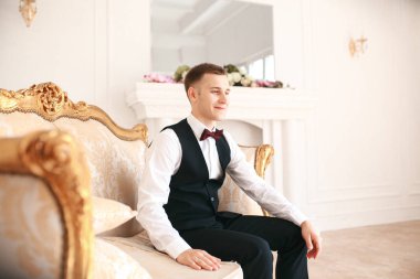 Damat kanepede onun düğününde gelin için bekleyen oturuyor. gülümseyen ve gelin için bekleyen düğün smokin. Zarif adam siyah kostüm ve papyon.