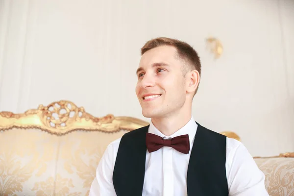 Sposo seduto sul divano in attesa della sposa il giorno del suo matrimonio. al matrimonio smoking sorridente e in attesa di sposa.Elegante uomo in costume nero e papillon . — Foto Stock