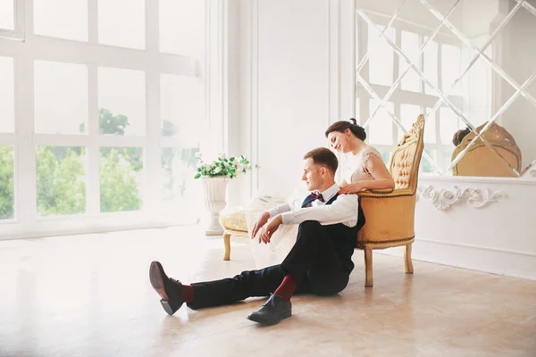Noiva em belo vestido e noivo em terno preto sentado no sofá dentro de casa em estúdio branco interior como em casa. Estilo de casamento na moda  . — Fotografia de Stock