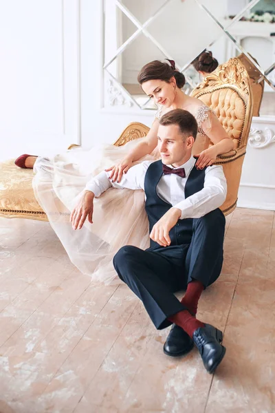 Novia en hermoso vestido y novio en traje negro sentado en el sofá interior en el interior del estudio blanco como en casa. Estilo de boda de moda  . — Foto de Stock