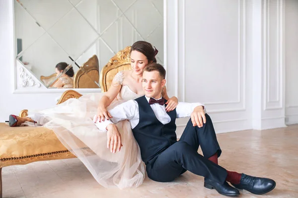 Novia en hermoso vestido y novio en traje negro sentado en el sofá interior en el interior del estudio blanco como en casa. Estilo de boda de moda  . — Foto de Stock