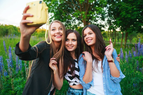 Tre hipsters flickor blondin och brunett tar självporträtt på polaroidkamera och leende utomhus. Flickor att ha roligt tillsammans i parken. — Stockfoto