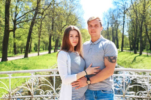 Pohledný muž a jeho krásná těhotná manželka jsou objímání a usmívá se přitom stát v parku — Stock fotografie
