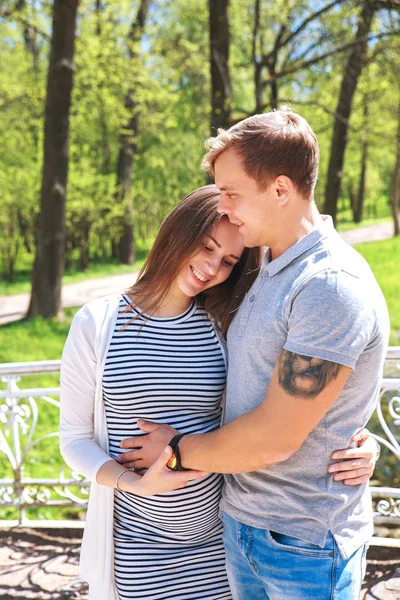 Bonito homem e sua bela esposa grávida estão abraçando e sorrindo enquanto estão de pé no parque — Fotografia de Stock