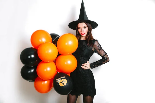 Portret van mooie jonge vrouw in zwarte heks halloween kostuum met oranje en zwarte ballonnen op witte achtergrond — Stockfoto