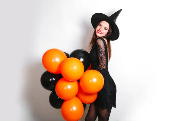 Portret van mooie jonge vrouw in zwarte heks halloween kostuum met oranje en zwarte ballonnen op witte achtergrond — Stockfoto