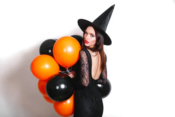 Portret van mooie jonge vrouw in zwarte heks halloween kostuum met oranje en zwarte ballonnen op witte achtergrond — Stockfoto