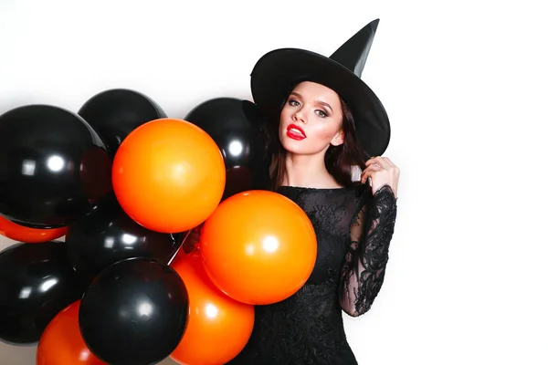 Portret van mooie jonge vrouw in zwarte heks halloween kostuum met oranje en zwarte ballonnen op witte achtergrond — Stockfoto