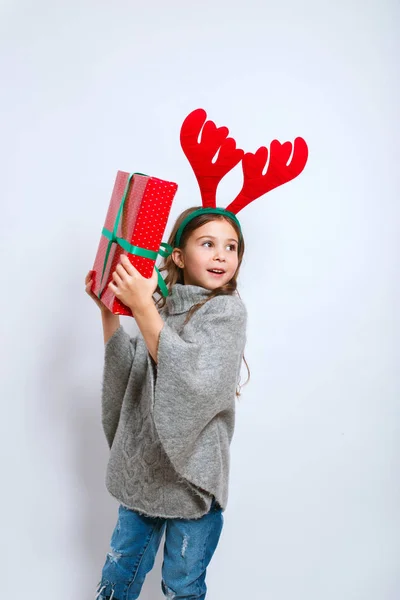 Счастливая маленькая улыбчивая девочка с подарочной коробкой на Рождество. Улыбающаяся смешная девушка в оленьих рогах в студии . — стоковое фото