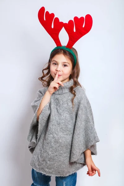 Счастливая маленькая улыбчивая девочка держит палец на губе секретной концепции Рождества. Улыбающаяся смешная девушка в оленьих рогах в студии . — стоковое фото