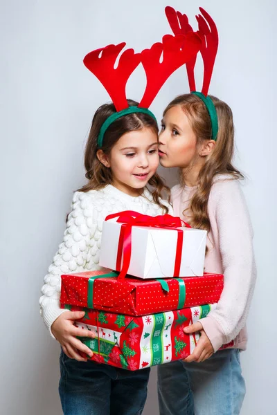 Две счастливые маленькие улыбающиеся девочки с рождественскими подарочными коробками... рождественская концепция. Улыбающиеся смешные сестры в оленьих рогах в студии рассказывают секрет . — стоковое фото