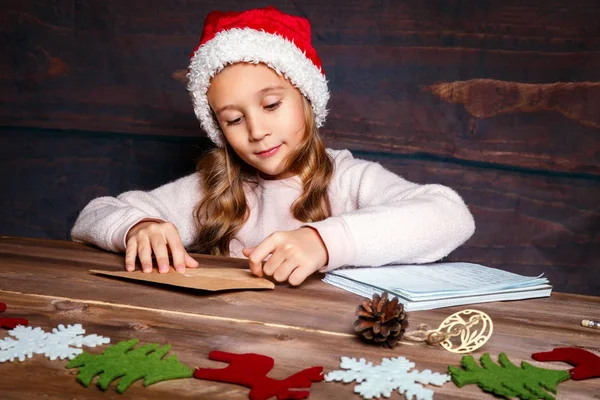 Ребенок пишет письмо Санта-Клаусу. Забавная девушка в шляпе Санты пишет Санте письмо. — стоковое фото