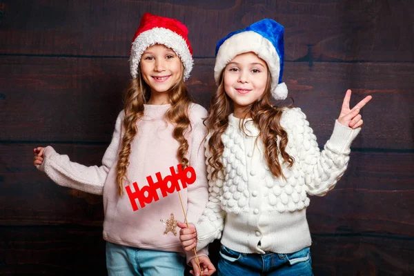 Две маленькие улыбающиеся девочки веселятся. Смешные сестры в шляпе Санты на деревянном фоне — стоковое фото