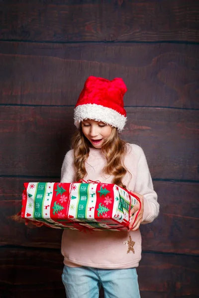 Счастливая маленькая улыбчивая девочка с подарочной коробкой на Рождество. Смешная девушка в шляпе Санты на деревянном фоне . — стоковое фото