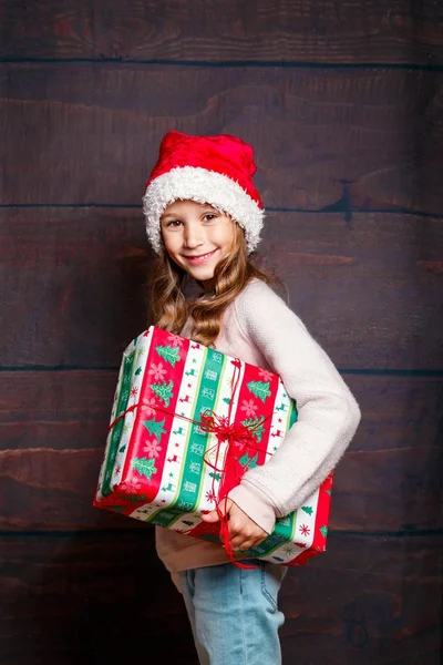 Счастливая маленькая улыбчивая девочка с подарочной коробкой на Рождество. Смешная девушка в шляпе Санты на деревянном фоне . — стоковое фото
