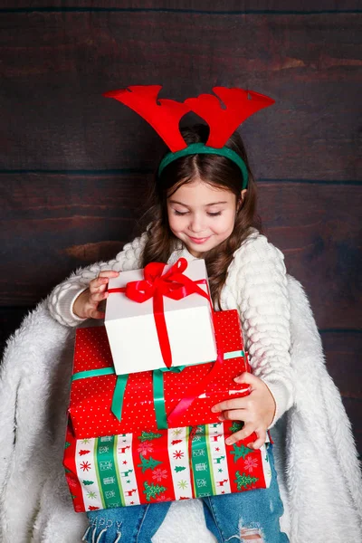 Feliz niña sonriente con cajas de regalo de Navidad.. concepto de Navidad. Sonriente niño divertido en cuernos de ciervo sobre fondo de madera —  Fotos de Stock
