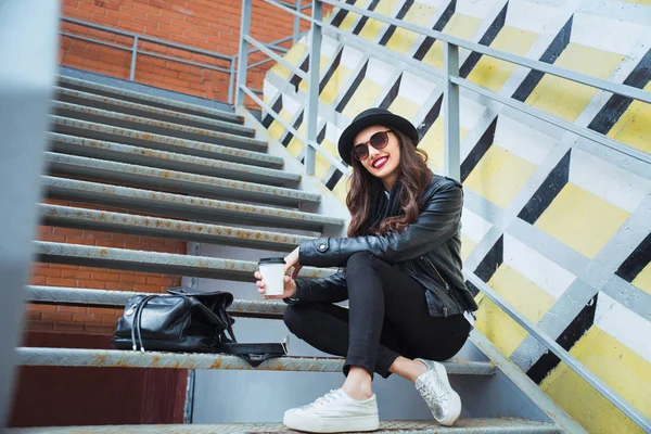 Молодая стильная женщина на городской улице сидит и пьет кофе или чай  . — стоковое фото