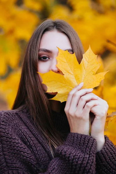 Beautiful young woman in a brown coat holding a bright yellow autumn leaf near the face in the park. — Stock Photo, Image