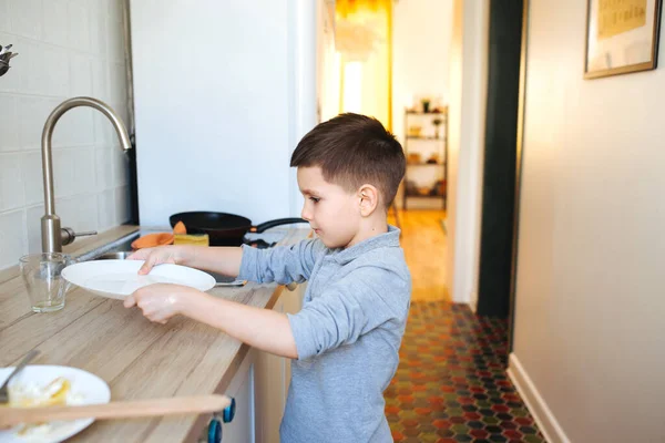 Kleiner Junge spült Geschirr in der heimischen Küche. Kind hilft seinen Eltern bei der Hausarbeit — Stockfoto