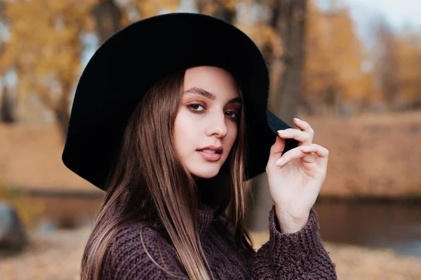 Zbliżenie portret pięknej dziewczyny w czarnym kapeluszu stojącej w pobliżu kolorowych jesiennych liści. — Zdjęcie stockowe