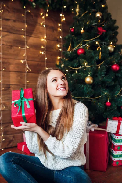 Gyönyörű lány, piros karácsonyi ajándékkal. Boldog nő Mikulás kalap ül közel újévi fa — Stock Fotó