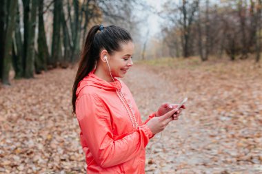 Kulaklıklı genç bir kadın sonbahar parkında koşuyor ve cep telefonu arıyor.