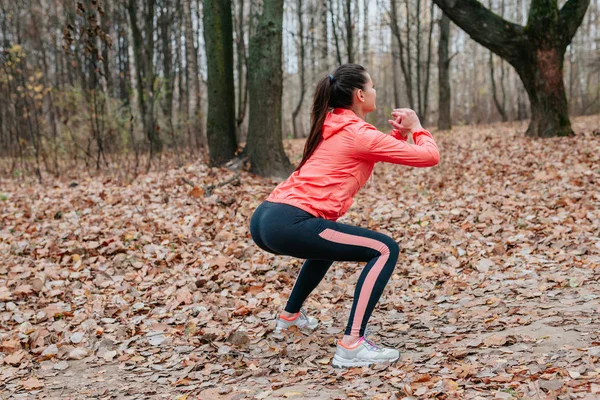 Young woman do squats outdoor on autumn leaves at the park.