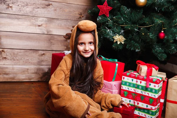 Забавная маленькая девочка в пижаме сидит с подарочной коробкой рядом с елкой — стоковое фото